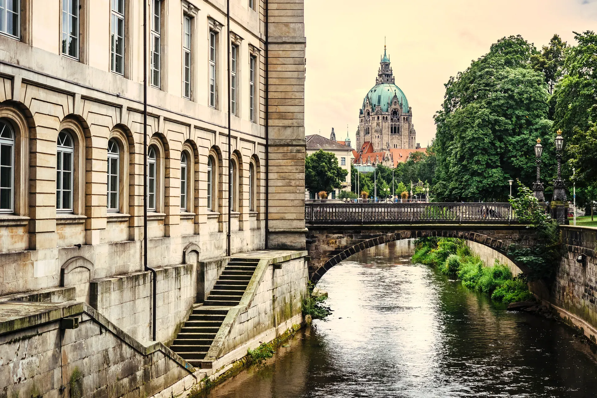 Blick über einen Fluss mit Steinbrücke auf die Kuppel des Neuen Rathauses in Hannover